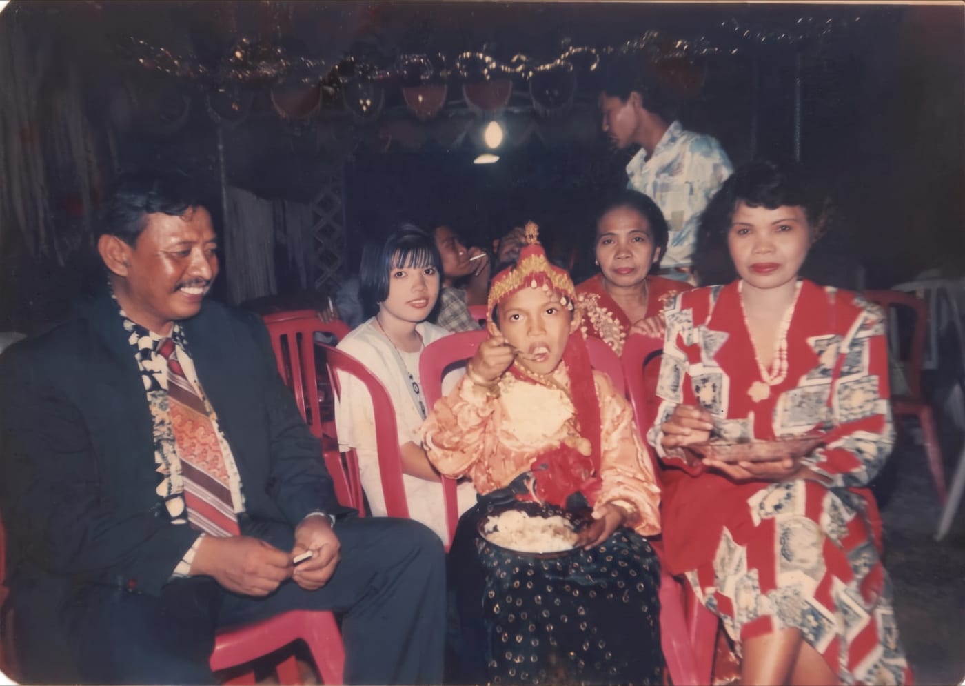 Restored colour photograph of a family at a ceremony, a child in a gold headdress being fed