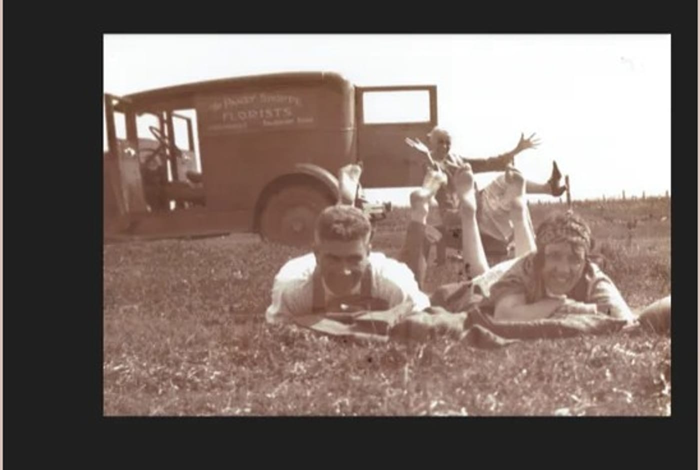 Restored photograph of young people lying on grass in front of a florist's delivery truck