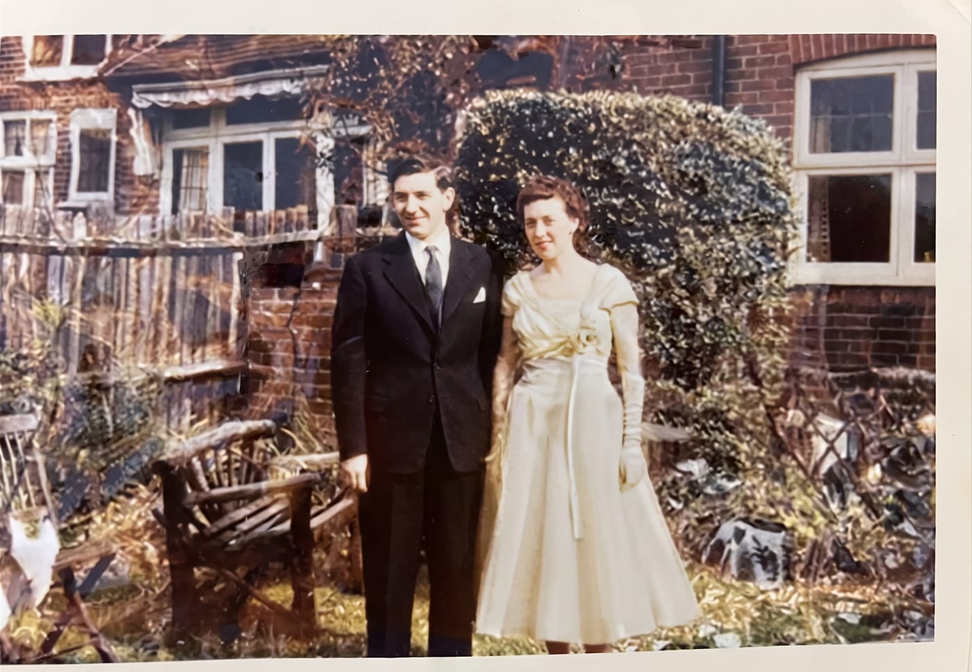 Restored colour photograph of a couple in wedding clothes standing in a garden in front of a brick house