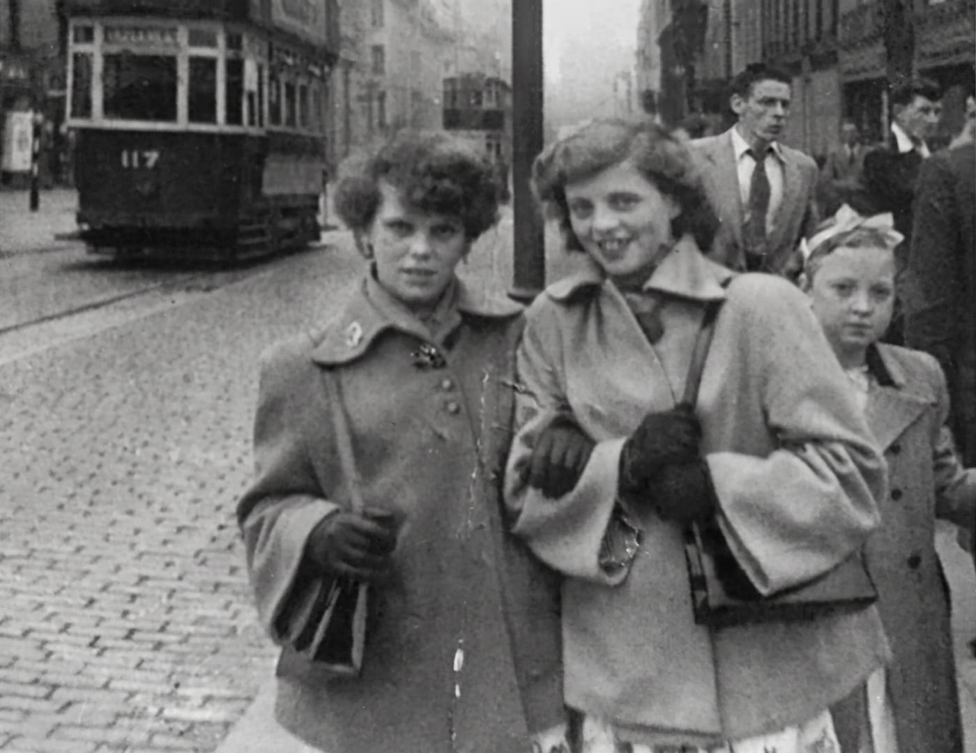Restored photograph of two women walking arm in arm on a cobbled street with a tram behind them